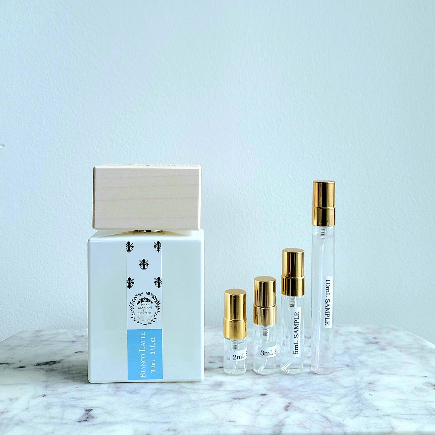 Bianco Latte Perfume samples, featuring a white square bottle with a natural wood cap and silver floral motifs, accompanied by four smaller samples in clear vials with gold caps, displayed on a marble surface